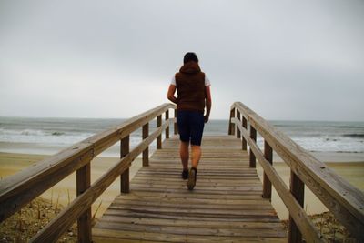 Full length rear view of person walking on pier at beach against sky