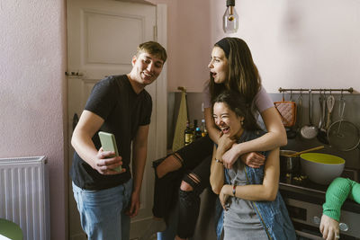 Smiling man sharing smart phone with cheerful female friends in kitchen at home