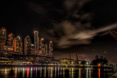 Illuminated modern buildings by river against sky at night