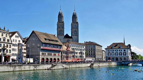 Grossmunster cathedral by limmat river in city against blue sky