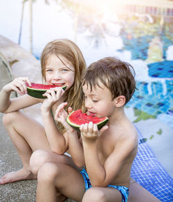 Siblings eating watermelon while sitting at swimming pool