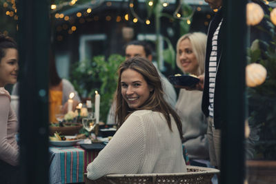 Smiling young woman looking back while sitting with friends in glass conservatory in back yard