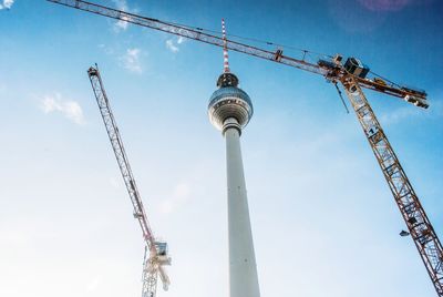 Low angle view of communications tower and cranes against blue sky during sunny day