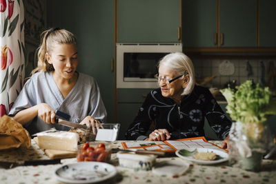 Female caregiver cutting bread loaf at dining table while sitting by senior woman in kitchen