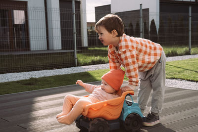 Brother giving a ride to baby sister in big toy car outdoors. children having fun near house. boy