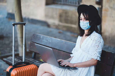 Beautiful woman flu mask using laptop sitting on bench outdoors