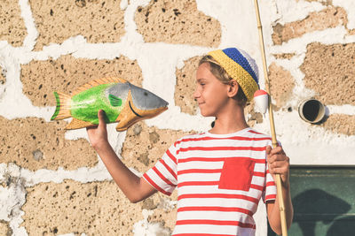 Smiling boy holding fish while standing against wall