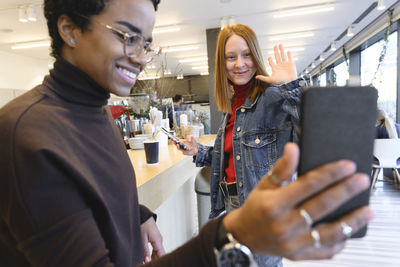 Woman waving hand at mobile phone while taking selfie with friend in cafe