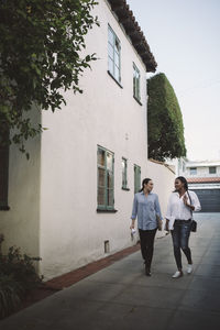 Full length of female designers discussing while walking on sidewalk by building against sky