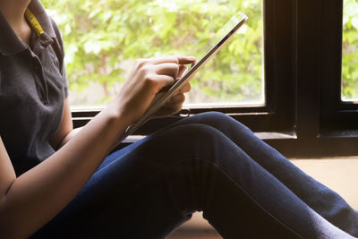 Midsection of woman using mobile phone while sitting on window