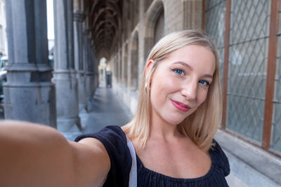 Lovely blonde woman in stylish clothes making selfie while walking past old building.