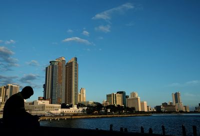 Man sitting at riverbank in city against blue sky