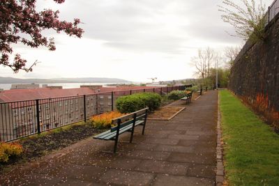 Empty park bench by footpath in city against sky