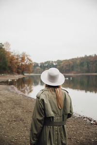 Rear view of woman wearing hat standing by lake against sky