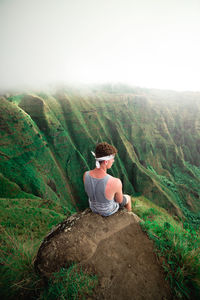 Rear view of man sitting on rock at cliff against sky