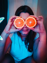 Mid adult woman holding orange fruit in front of eyes while sitting against wall