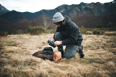 Side view of man in hat and warm jacket taking accessories from camera in meadow with dry grass