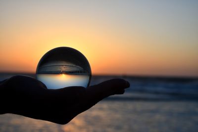 Cropped hand of person holding crystal ball against sea and sky during sunset