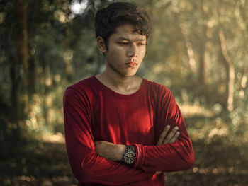 Young man with arms crossed looking away while standing against trees in forest