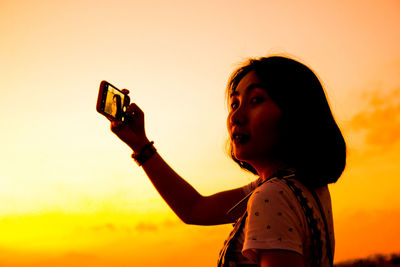 Portrait of young woman photographing with smart phone against sky during sunset