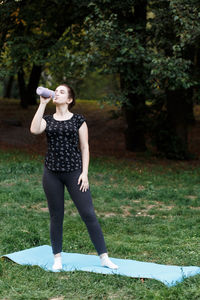 The relaxed girl is doing yoga in the park on carpet and is drinking w