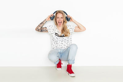 Portrait of happy woman holding headphones while crouching against wall