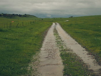 Dirt road amidst grassy field against cloudy sky