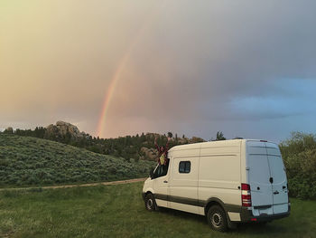 Happy woman with arms raised peeking through window of motor home in forest against cloudy sky during sunset