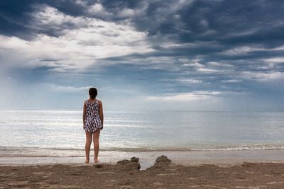 Rear view of woman standing on beach against cloudy sky
