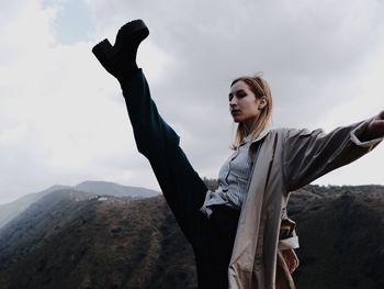 Woman kicking while standing on mountain against sky