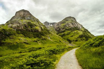 Narrow road leading towards mountains against clouds