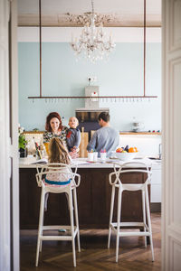 Smiling woman carrying son while looking at girl studying in kitchen at home