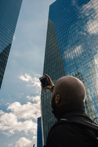 Low angle view of man taking selfie on mobile phone against modern buildings and sky