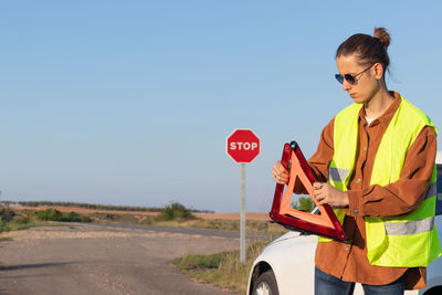 Man on sunglasses setting down red warning triangle in the middle of the road and stop sign