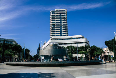 People sitting by fountain in city against sky