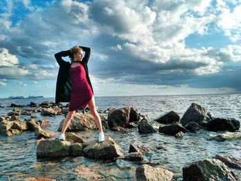 Young woman posing on rocks in sea against cloudy sky