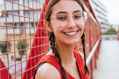 Delighted female in stylish glasses and with pigtails standing near net fence in street and looking at camera