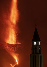 Dramatic scenery of church clock tower silhouette on mountain slope against powerful eruption of cumbre vieja volcano at night in la palma island