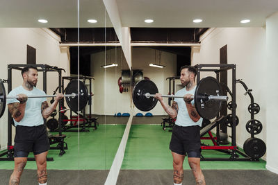 Sportsman lifting barbell while standing by mirror in gym