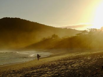 Scenic view of sea against sky during sunset with a surfer walking at the sand