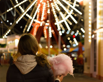 Rear view of woman holding cotton candy against illuminated ferris wheel at night