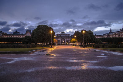 Illuminated road by buildings against sky at dusk