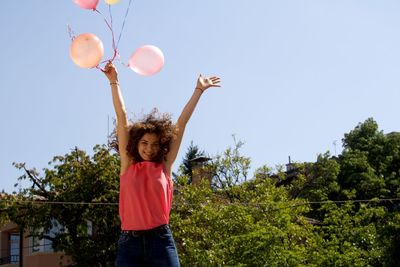Happy young woman with balloon jumping against sky during sunny day