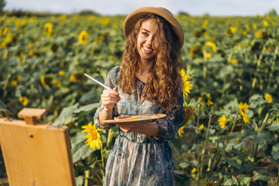 Young woman holding sunflower while standing against yellow flowers