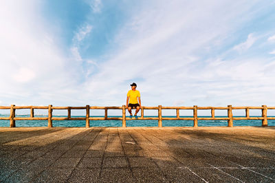 Man sitting on railing at promenade against cloudy sky