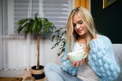An attractive blonde is sitting on the sofa with a mug of coffee.