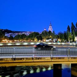 Bridge over river by illuminated buildings against blue sky