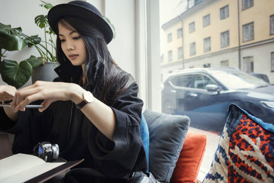 Young female blogger photographing through smart phone while sitting by window in office