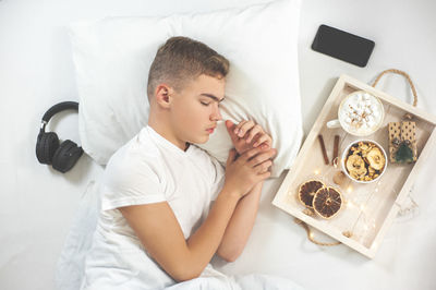 A teenage boy in a white t-shirt sleeps in bed with a smartphone and headphones. 