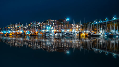 Sailboats moored on illuminated harbor by buildings against sky at night